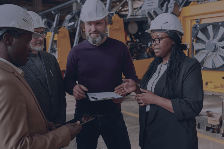 Four construction workers, three men and one woman, in hard hats engaged in conversation on a job site.