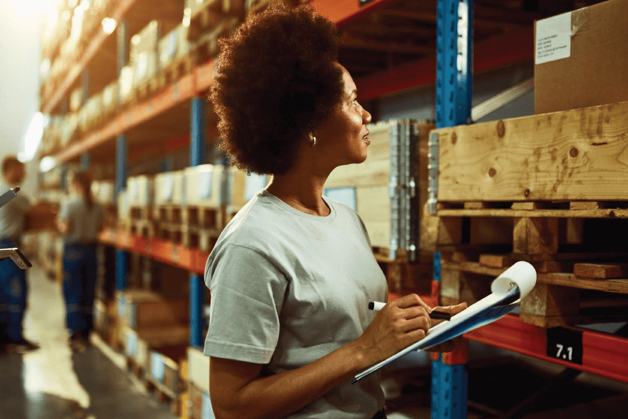 A woman stands in a warehouse holding a clipboard, surveying the space with a focused expression.